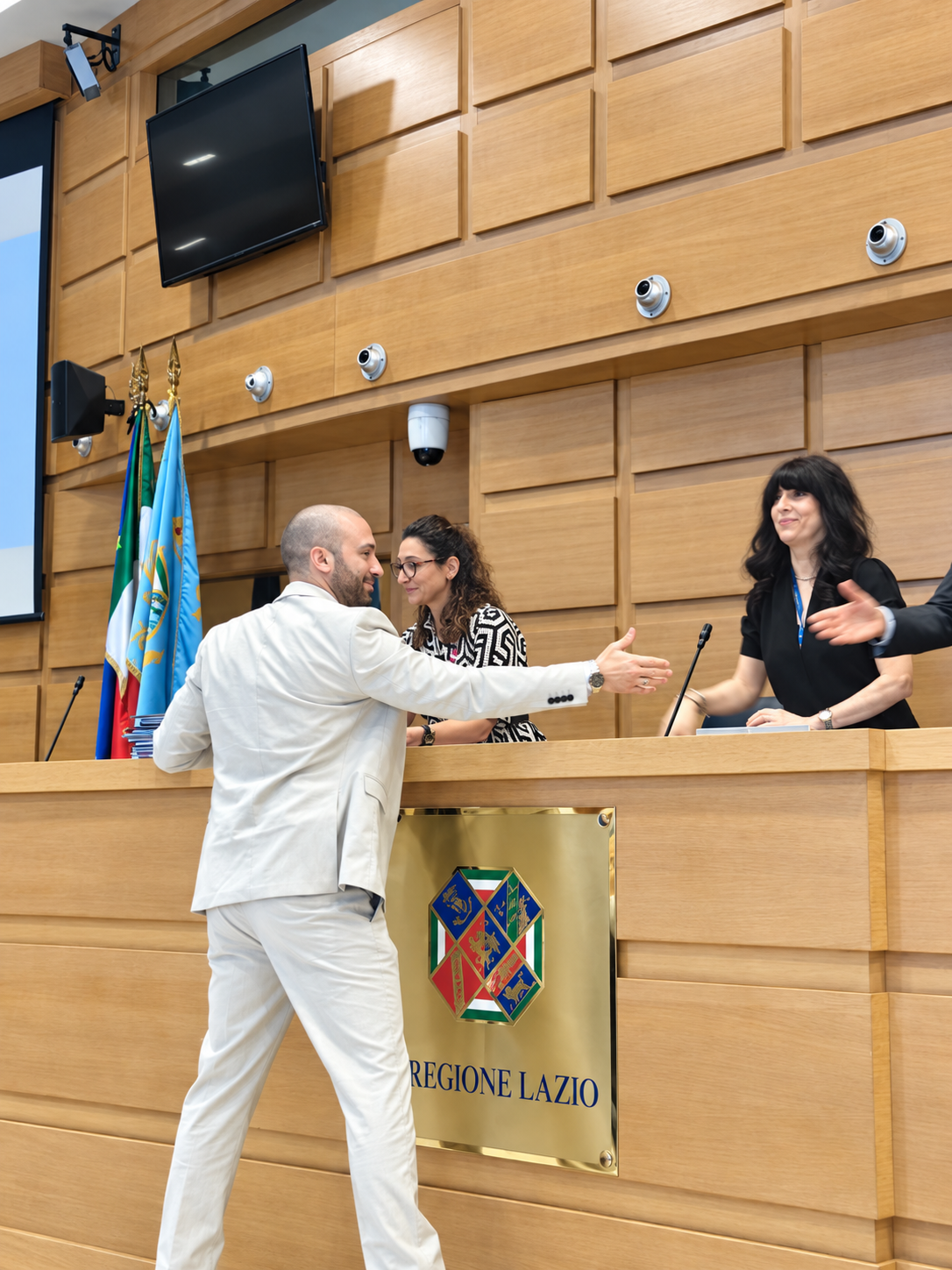 Un uomo in abito chiaro saluta alcune persone dietro un banco con lo stemma della Regione Lazio.